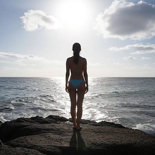 Silhouetted woman in blue bikini stands on rocky shore, gazing at sunlit ocean, with bright sky and scattered clouds. Photograph.
