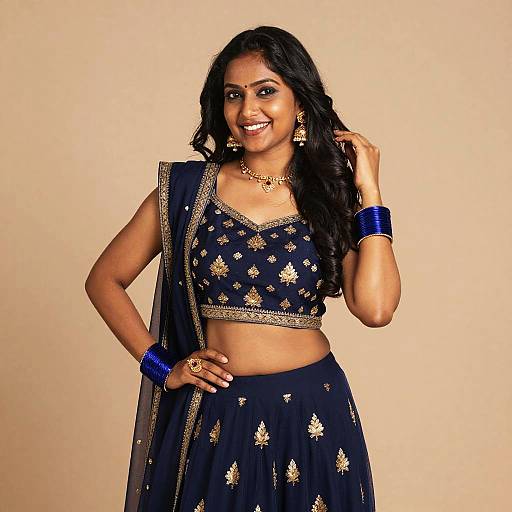 Photograph of a smiling Indian woman with dark hair, wearing a navy blue, gold-embroidered crop top and skirt, with matching blue b