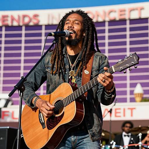 Photograph of a bearded black man with dreadlocks, wearing a denim jacket and playing an acoustic guitar, singing into a microphone on stage with a