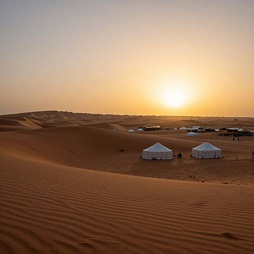 Photograph of a desert sunset with golden sky, orange sun, white tents scattered across rippled sand dunes, and distant camp structures.