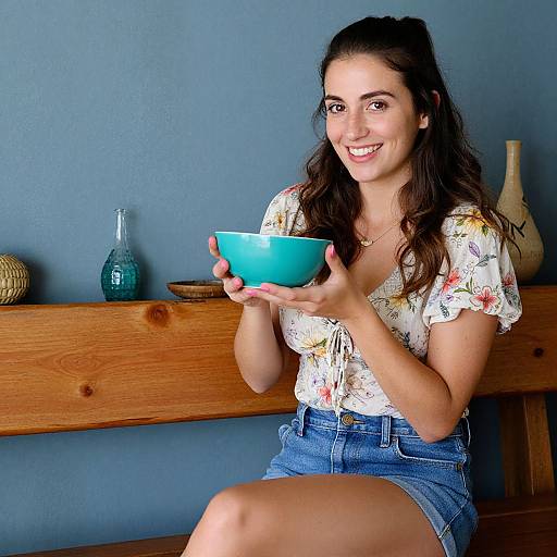 Photograph of a smiling young woman with long brown hair, wearing a floral shirt and denim shorts, holding a blue bowl, sitting against a blue wall