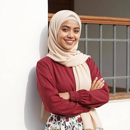 Photograph of a smiling young woman with light brown skin, wearing a white hijab, maroon long-sleeve blouse, and floral skirt,
