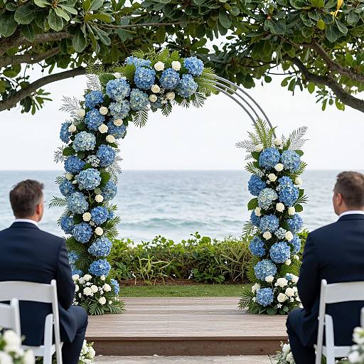 Photograph of two men in black suits, seated, facing a blue and white floral arch overlooking a calm sea and lush greenery.