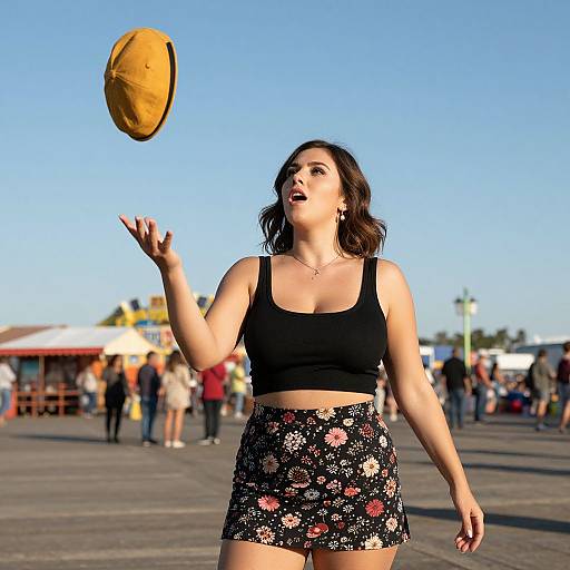 Photograph of a young woman with medium skin tone, dark wavy hair, wearing a black crop top and floral skirt, catching a brown hat in