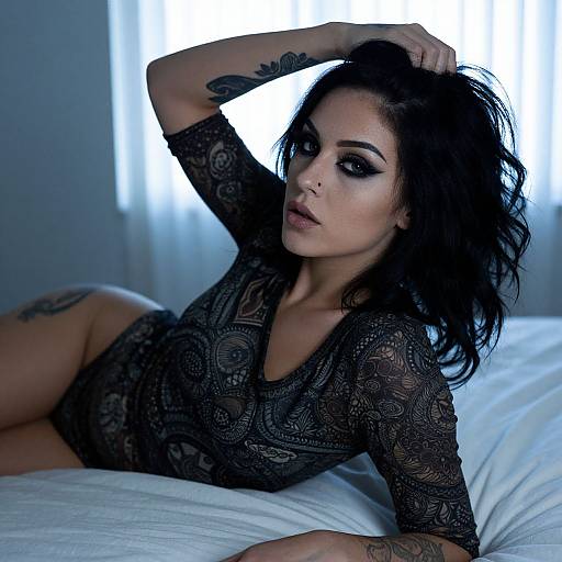 Photograph of a dark-haired woman with tattoos, wearing a black lace bodysuit, lying on a white bed, hand in hair, against a