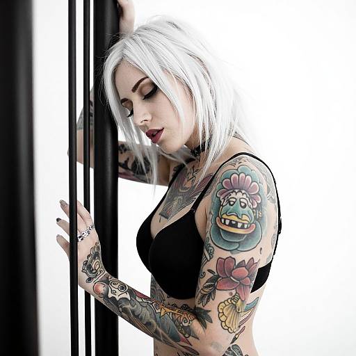 Photograph of a white-haired, tattooed woman with colorful arm tattoos, wearing a black bra, leaning against a black vertical bar.