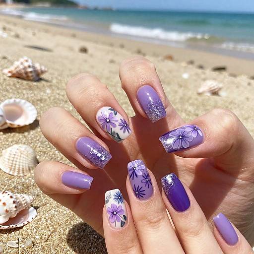 Photograph of a hand with purple and floral-accented nails on a sandy beach with scattered seashells and ocean in the background.