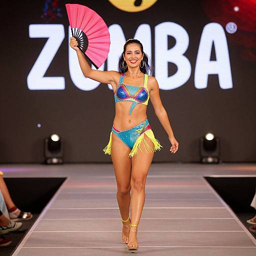 Photograph of a confident woman in a colorful, beaded bikini with yellow fringe, holding a pink fan, walking a runway with 