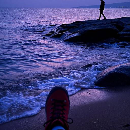 Photograph of a beach at twilight, featuring silhouetted waves, a person walking on rocks, and a red shoe in the foreground.