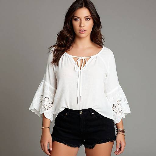 Photograph of a young woman with long dark hair, wearing a white lace-trimmed blouse and black denim shorts, standing against a gray background.