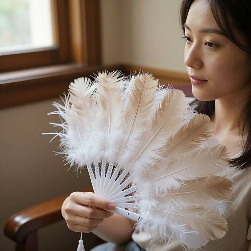Photograph of an Asian woman with dark hair holding a white feather fan, illuminated by soft natural light, in a wooden-walled room.