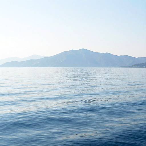 Photograph of a calm, blue ocean with gentle ripples in the foreground, leading to a distant, hazy mountain range under a bright, clear
