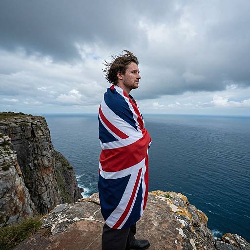 Photograph of a man with messy brown hair, standing on a cliff, wrapped in the British flag, overlooking a stormy ocean.