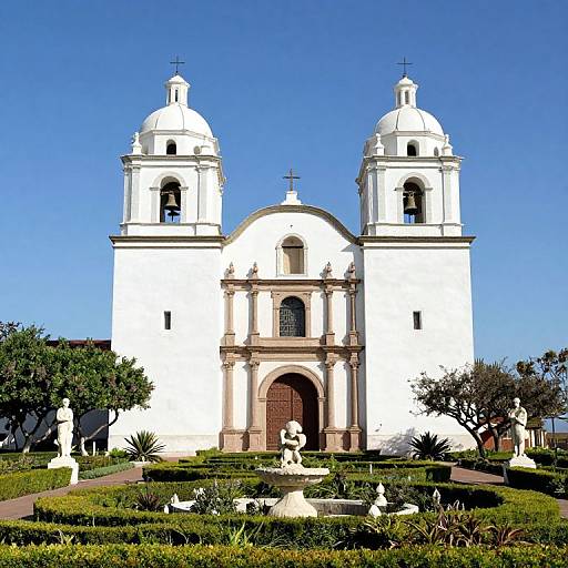 Photograph of a white, twin-dome Spanish colonial church with arched doorway, surrounded by manicured gardens, statues, and clear blue sky.