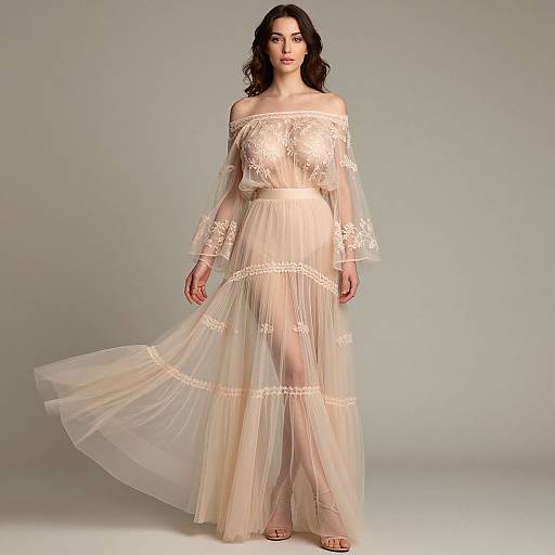 Photograph of a young woman with long dark hair in an off-shoulder, sheer, lace-trimmed, beige maxi dress, standing against