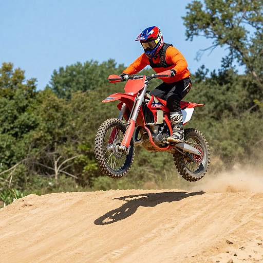 Photograph of a motocross rider in bright orange gear and red helmet, mid-air on a sandy hill, surrounded by green trees.