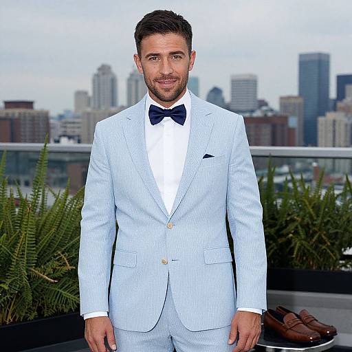 Photograph of a handsome man with short dark hair and beard, wearing a light blue pinstripe suit, white shirt, black bow tie, standing