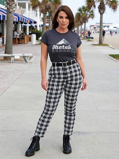 Photograph of a fit, brunette woman in a black t-shirt and black-and-white plaid pants, standing on a sunny sidewalk with palm trees and