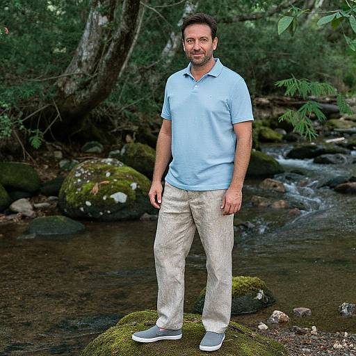 Photograph of a middle-aged man with short brown hair and beard, wearing a light blue polo shirt and beige pants, standing on a mossy rock