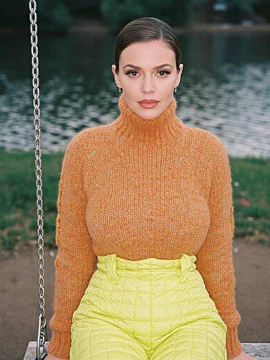 Photograph of a fair-skinned woman with dark hair in an orange turtleneck sweater and high-waisted yellow pants, sitting on a swing