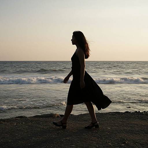 Silhouetted woman in a flowing dress walks on a pebbled beach at sunset, waves gently crashing in the background. Photograph.