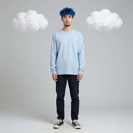 Photograph of a young man with blue spiky hair, wearing a light blue sweatshirt, black pants, and black sneakers, standing against a gray