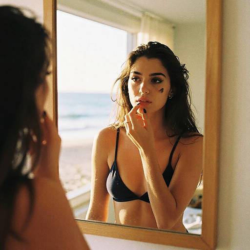 Photograph of a dark-haired woman in a black bra, standing in front of a mirror, softly touching her lips, with sunlight streaming in from a
