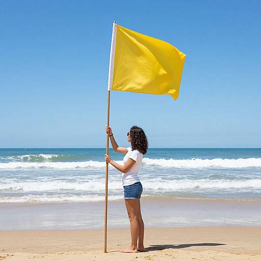 Photograph of a curly-haired woman in white shirt and denim shorts, holding a yellow flag on a sunny beach with blue sky and ocean waves in the
