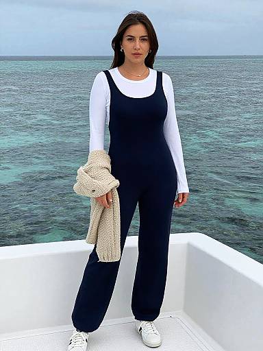 Photograph of a woman with medium skin tone, dark hair, wearing a black jumpsuit over a white long-sleeve shirt, holding a kn