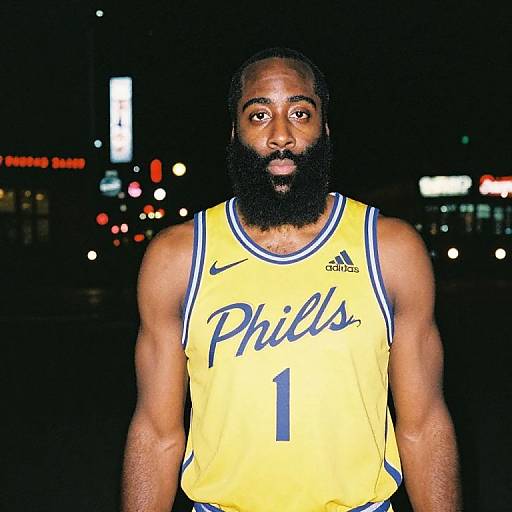 Photograph of a muscular Black man with a full beard, wearing a yellow Philadelphia 76ers basketball jersey with number 1, standing at night with