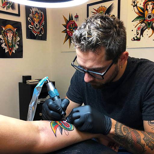 Photograph of a tattoo artist with spiky black and gray hair, glasses, and tattoos, meticulously tattooing a colorful design on a person's arm