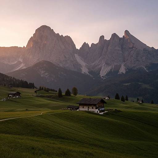 Photograph of a serene alpine landscape at sunset, featuring lush green hills, rustic chalets, and towering jagged mountains under a soft pink and