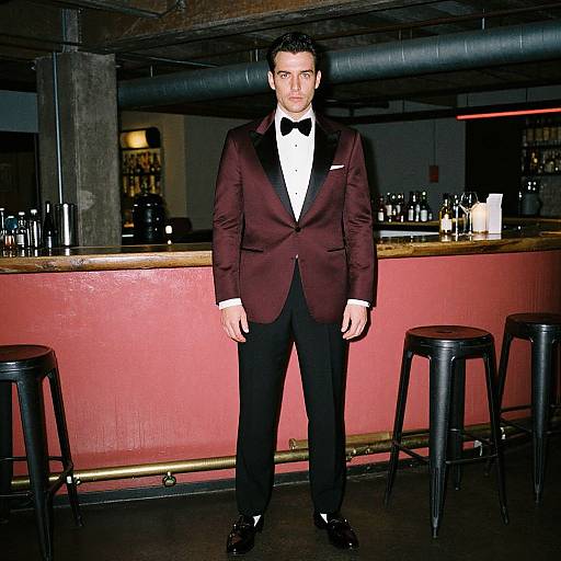 Photograph of a tall, serious-looking man in a dark burgundy tuxedo with black bow tie, standing in a dimly lit bar with