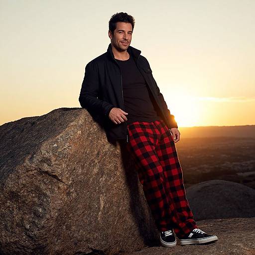 Photograph of a bearded man in black jacket, black shirt, red plaid pants, and black sneakers, leaning against a large rock at sunset