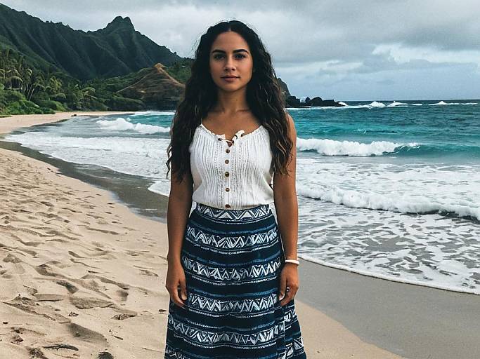 Photograph of a young woman with long curly hair, wearing a white blouse and blue patterned skirt, standing on a sandy beach with waves and mountains