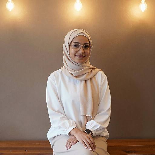 Photograph of a smiling Muslim woman with glasses, beige hijab, and white long-sleeve shirt, seated against a warm-lit, gray