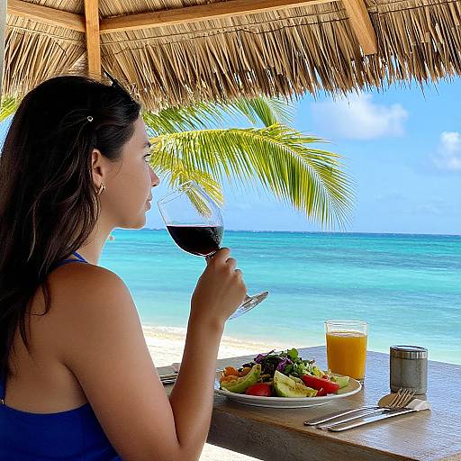 Photograph: Asian woman with dark hair sipping red wine at beachside bar, tropical palm, ocean view, colorful salad, orange juice, under