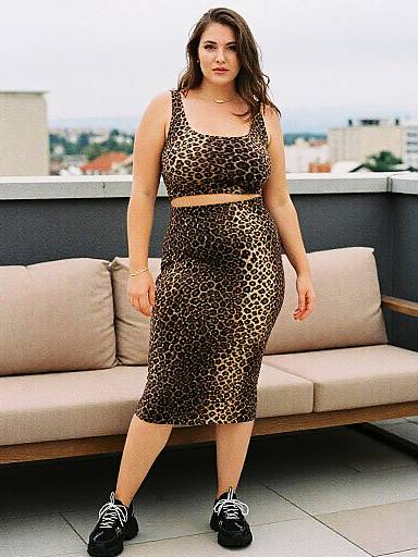 Photograph of a curvy woman with long brown hair in leopard print crop top and skirt, black sneakers, standing on balcony.