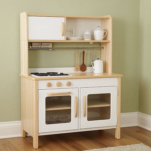 Photograph of a wooden and white children's play kitchen with stove, oven, shelves, utensils, and a white kettle on green wall.