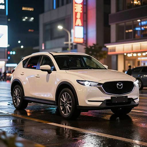 Photograph of a sleek white Mazda SUV driving on a wet, neon-lit city street at night, with reflections on the road.