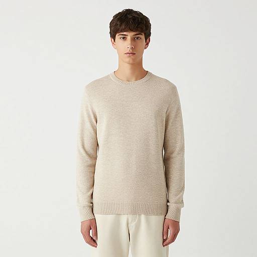 Photograph of a young man with short dark hair, wearing a beige knit sweater and white pants, standing against a plain white background.