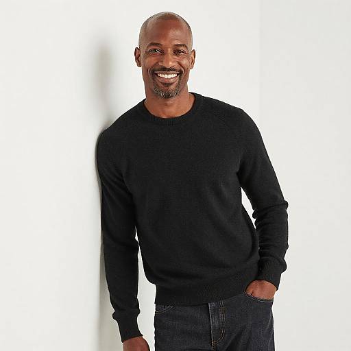 Photograph of a smiling, bald Black man with a goatee, wearing a black sweater and dark jeans, standing against a white background.