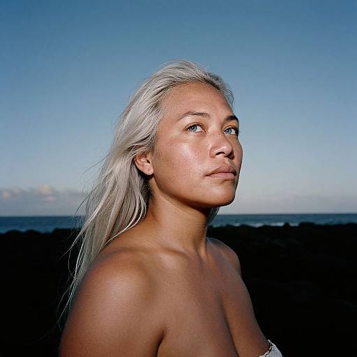 Photograph of a topless, tan-skinned woman with long, silver hair, gazing into the distance against a clear blue sky and dark ocean