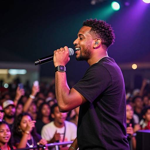 Photograph of a Black male singer with curly hair, beard, black t-shirt, and wristwatch, passionately performing into a microphone on stage, with