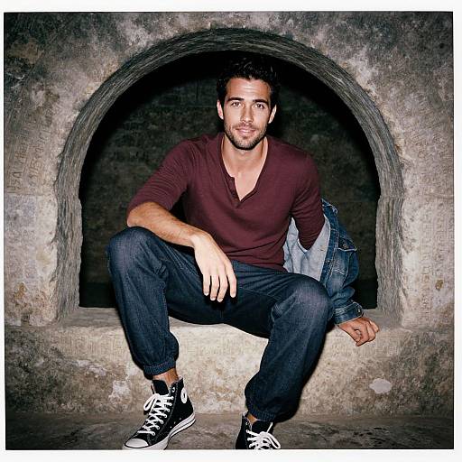 Photograph of a handsome, bearded man with dark hair, wearing a burgundy V-neck shirt, black jeans, and black Converse sneakers,