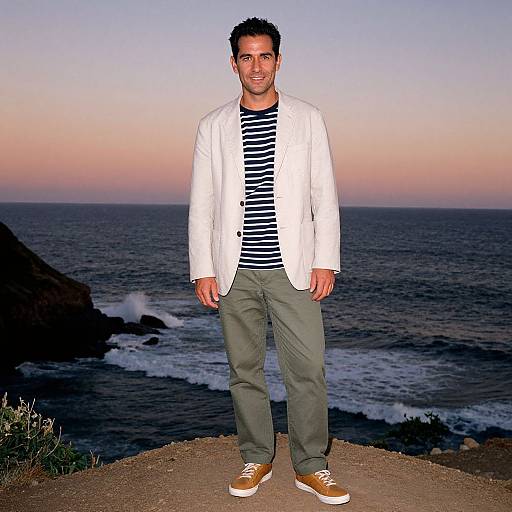 Photograph of a smiling man with short dark hair, wearing a white blazer, black-and-white striped shirt, green pants, and brown shoes,