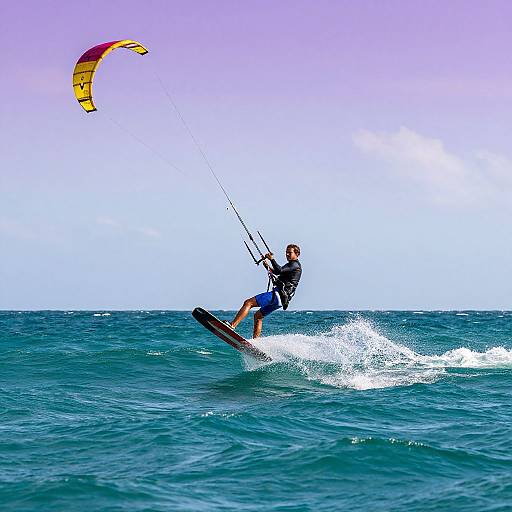 Photograph of a male kite surfer in blue shorts and black top, riding a wave, with a colorful kite in the sky.