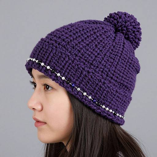 Photograph of a young girl with light brown skin and dark hair wearing a purple crocheted hat with a pom-pom, white bead trim,