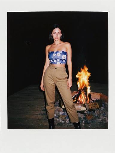 Photograph of a woman with dark hair, wearing a blue and white patterned strapless top and tan high-waisted pants, standing in front