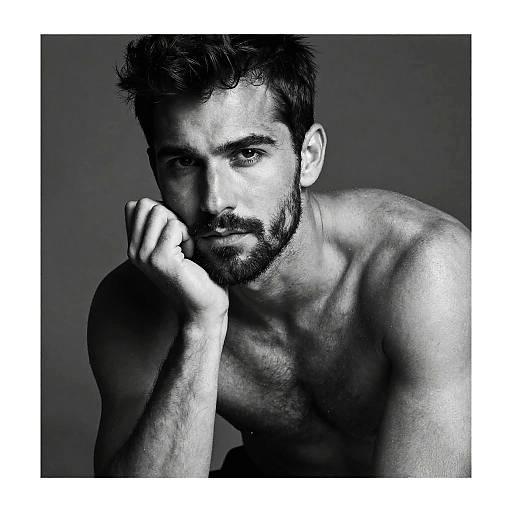 Black-and-white photograph of a shirtless, muscular man with short, curly hair, beard, and intense gaze, resting his chin on his hand against